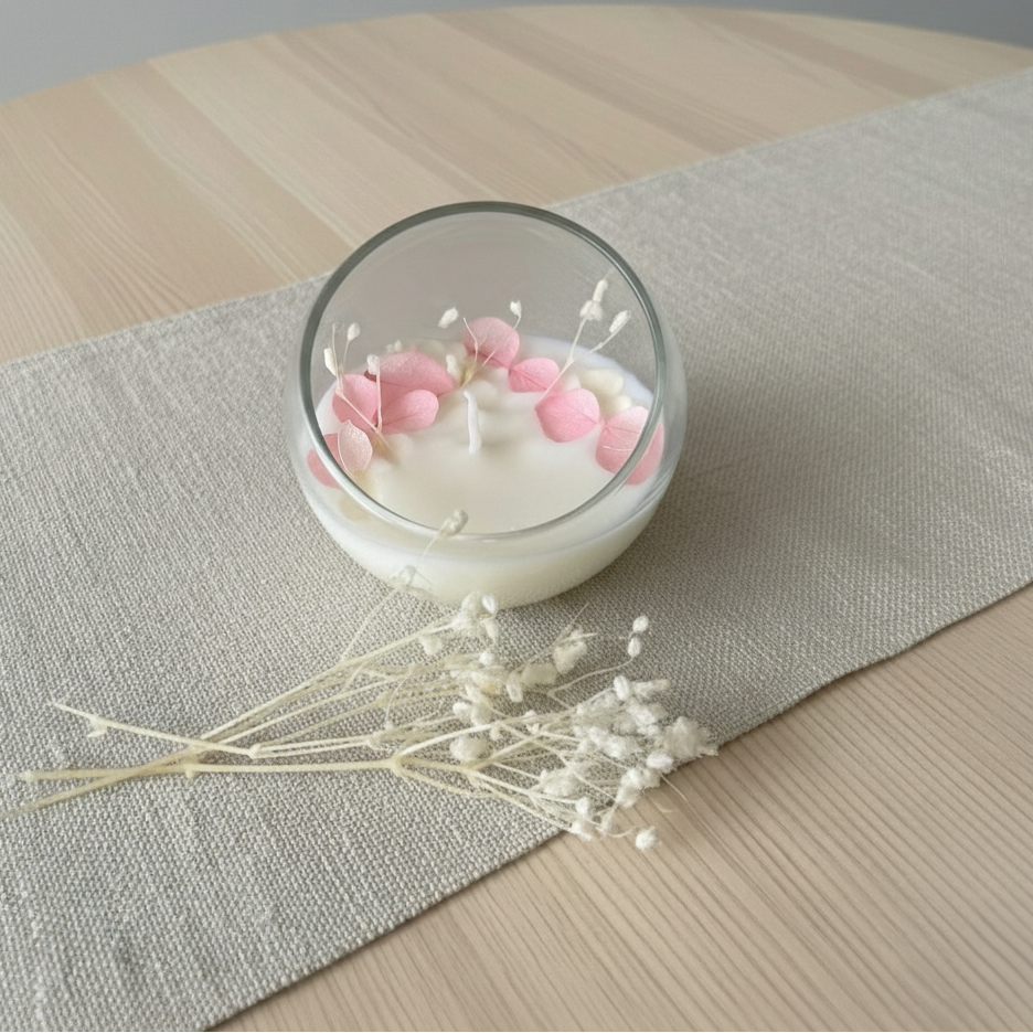 Candela decorativa artigianale in cera di soia con fiori essiccati rosa in contenitore in vetro sferico.
